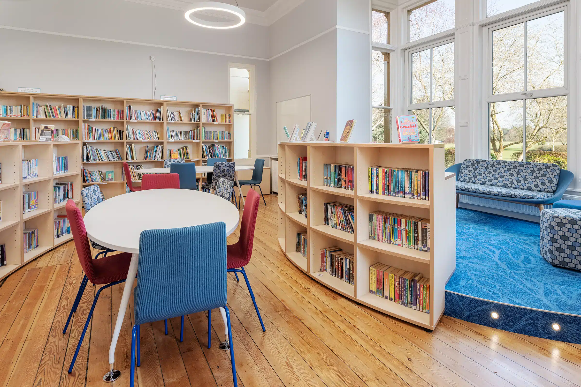 A bright school library with curved bookshelves, round tables, colourful chairs and a seating area by large windows.